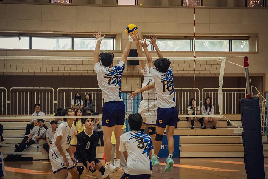 Two high-school-age players leaping together to block a ball at the net during a gym match